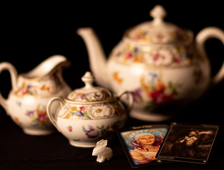 Tea pot, sugar bowl, and creamer on black background with small ivory rabbit and two tarot cards.